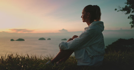 Young woman is enjoying a peaceful moment at sunset while sitting on a grassy hilltop overlooking a...