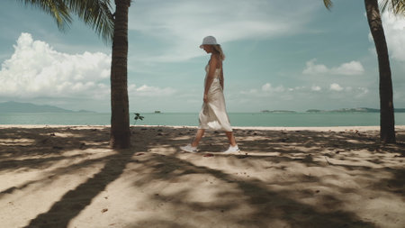 Elegant girl in white dress and hat walks exotic sand beach in Thailand, palm trees and turquoise water in background. Tropical island travel, outdoor lifestyle, enjoy summer holiday vacation, relaxの写真素材