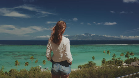 Young woman is taking in the breathtaking view of a tropical island paradise, with its crystal-clear turquoise waters, lush greenery, and majestic mountains in the distanceの写真素材