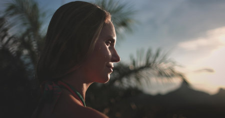 Young woman is smiling while enjoying a beautiful sunset in a tropical location with palm trees and mountains in the backgroundの写真素材