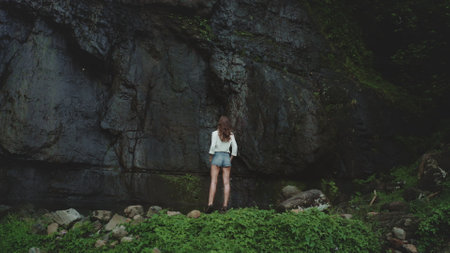 Young woman stands near the bottom of a jungle cliff face, taking in the natural beauty of a tropical island during the rainy seasonの写真素材