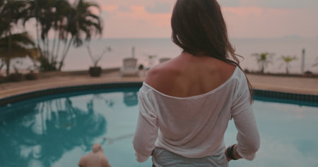 Woman on tropical vacation walking resort hotel area at evening. Tourist woman enjoy swimming pool and ocean seascape in pink sunset light. Outdoor lifestyle travel on summer. Back viewの写真素材