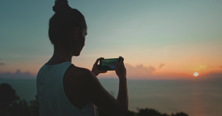 Capturing the sunset at the beach with a smartphone, a woman enjoys the colorful sky reflecting on the ocean, creating lasting memories of her travelsの写真素材