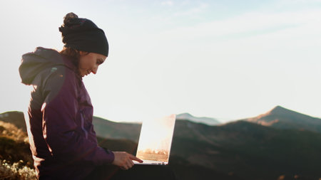 Woman working on a laptop in the mountains, surrounded by nature. Enjoying freedom and adventure, she types away, connecting with the worldの写真素材