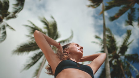 Young sportswoman is tying her hair after a workout on a tropical beach. She is enjoying a moment of relaxation surrounded by palm treesの写真素材