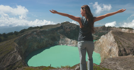Woman stands with arms outstretched on mountain summit Volcanic Kelimutu blue crater lake. Travel destination. Beautiful wild landscape. Female tourist on summer vacation. Lifestyle holiday. Back viewの写真素材