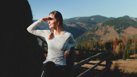 Young woman is sitting on a fence, shielding her eyes from the sun and enjoying the view of the mountains in the distanceの写真素材