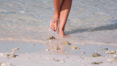 A person is walking along the sandy beach with their feet immersed in the gentle waves of the ocean. The individual appears to be enjoying the tranquil stroll beside the seashore.の写真素材
