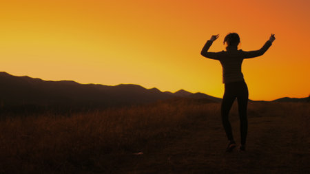 Girl dancing against bright orange sunset sky. Slim young girl silhouette having fun, moving, raise hands up, listening music in mountain field. Back view. Slow motion 4Kの写真素材