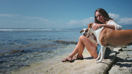 Young woman is sitting on a rock by the ocean, petting her two dogs on a sunny day. The dogs are enjoying the attention and the beautiful sceneryの写真素材
