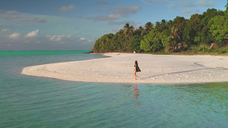 Young woman strolls on a sunny beach in french polynesia, capturing the tranquil paradise with a droneの写真素材