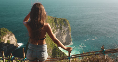 Young woman is taking in the beauty of the turquoise ocean and the dramatic cliffs of kelingking beach from a scenic viewpointの写真素材
