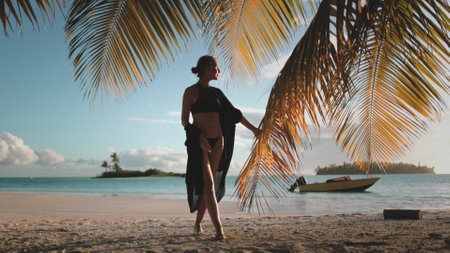 Perfect summer vacation background. Woman walk under palm tree on sunset sand beach. Barefoot female in black shirt touch coconut leaf enjoy relax travel holiday. Tropical wild island sea landscape.の写真素材