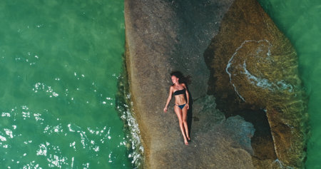 A person lounges on a large rock surrounded by turquoise water, soaking up the sun on a clear day. Waves gently lap against the shore, creating a serene atmosphere.の写真素材