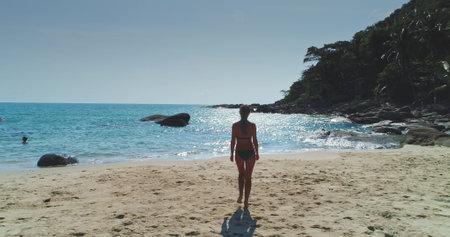 A woman strolls on a sandy beach, surrounded by rocks and clear water, with lush trees in the background under a sunny sky. Waves gently lap at the shore as she enjoys the serene landscape.の写真素材