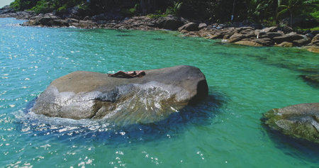A person lies comfortably on a large rock, soaking up the sun undisturbed, surrounded by crystal-clear turquoise water at a serene tropical beach. Lush greenery adds to the picturesque backdrop.の写真素材