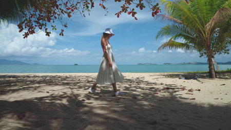 Young woman in a flowing dress and hat strolls leisurely on a sunny beach in thailand, surrounded by palm trees and ocean waves, creating a serene and peaceful atmosphereの写真素材