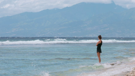 Young woman is wading in the ocean, enjoying the view of the waves crashing on the shore with mountains in the distanceの写真素材