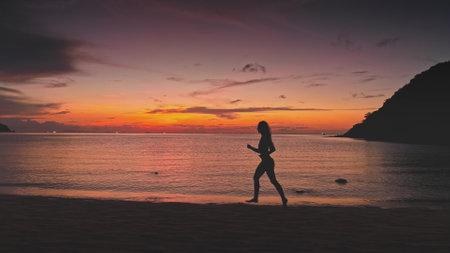Silhouette of sporty woman running barefoot on the beach in Thailand, enjoying stunning sunset over calm sea with colorful sky and distant islandsの写真素材