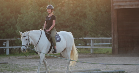 Horseback riding training, young jockey riding white horse in paddock near stables at sunset, equestrian sport, active lifestyle, love for horses, enjoying leisure time in natureの写真素材