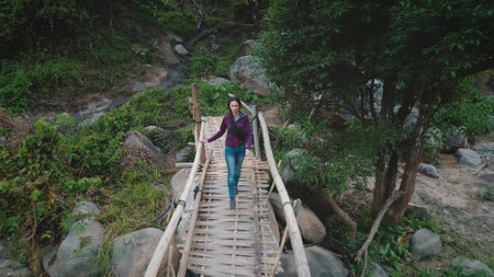 Aerial drone footage of a female tourist walking on a bamboo bridge across a small stream in a tropical forest, enjoying the tranquility of natureの写真素材