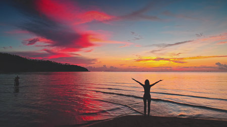 Silhouette of young woman standing on a tropical beach in Thailand, raising her arms and enjoying a beautiful colorful sunset over the ocean, reflecting red and orange colors on the waterの写真素材