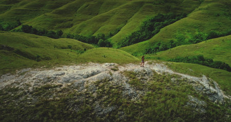 Aerial view of a young female tourist walking on a rocky cliff overlooking the rolling green hills...