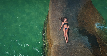 A person relaxes on a large, sunlit rock by the shoreline, surrounded by clear turquoise water. The...