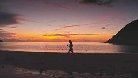 Silhouetted sportswoman running along a picturesque beach in Thailand, embracing the vibrant colors of a stunning sunset while celebrating freedom and a healthy lifestyleの写真素材