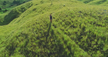 Aerial view capturing a female hiker traversing lush, grassy hills on Sumba Island, Indonesia, embracing the stunning landscape during a summer vacation filled with adventureの写真素材