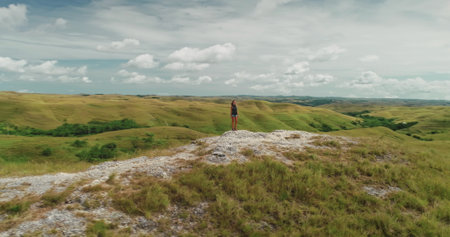 Young woman is standing on a rock admiring the breathtaking landscape of rolling green hills and cloudy sky in Sumba Island, Indonesia, embodying a sense of freedom and adventureの写真素材