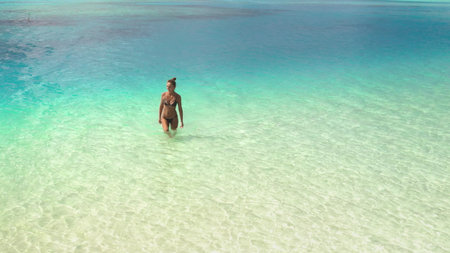 Young woman happily walks in crystal clear water of a tropical lagoon on a sunny day, enjoying the serene paradise of turquoise sea and white sandy beachの写真素材