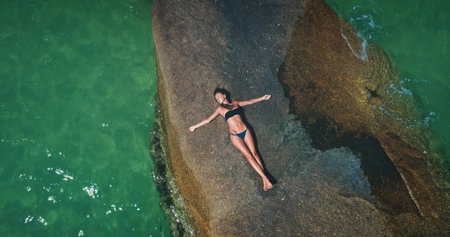 A woman lies on a flat rock surface surrounded by clear turquoise water. Sunlight enhances the vibrant colors as she enjoys a peaceful moment in a tropical beach setting.の写真素材