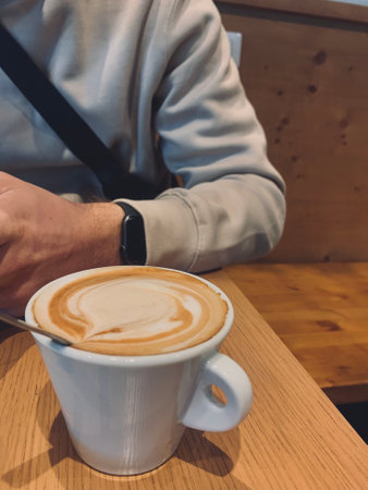 A man's hand with a clock holds coffee. Cappuccino in a coffee shop. Coffee cup on wood table in...