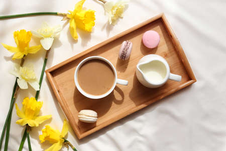Cozy home interior. Cup of coffee with milk and French macaroons on a wooden tray and yellow daffodil flowers. Breakfast in bedの写真素材