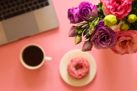 Girly workspace with flowers, laptop, donut, and coffee on pink background. Morning and breakfast...