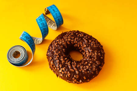 Concept of overweight and weight control. Unhealthy food. Chocolate glazed donut with sprinkles and a measuring tape, metric rules close-up on a bright yellow background. Flat lay, top view.の写真素材