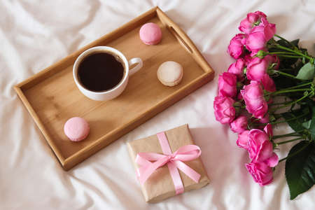Cozy holiday morning composition with roses, gift box and cup of coffee with macaroons on wooden tray. Mother's Day concept.の写真素材