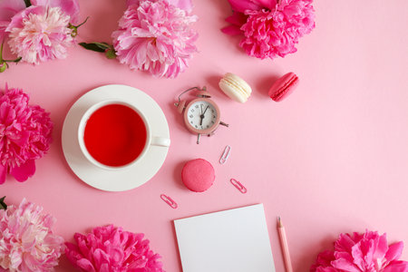 Beautuful flat lay with notepad, pencil, alarm clock, peony flowers, a cup of tea and macarons on a light pink background. Business, freelance and education concept. Morning time. Top viewの写真素材