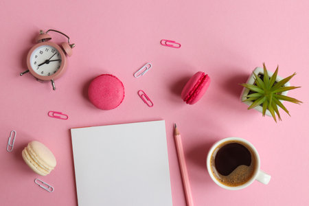 Workplace with notepad, pencil, alarm clock, plant, a cup of coffee and macarons on a pink background. Business, freelance and education concept. Morning time. Top view, flat lay.の写真素材