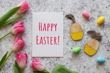 Easter greeting card. Composition with pink tulips, wooden chicken toys, colorful eggs and paper with text HAPPY EASTER against grey textured background. Top viewの写真素材