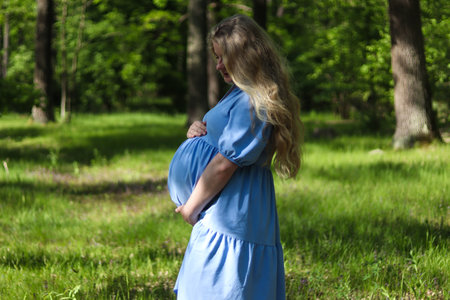 Beautiful young pregnant woman holding her belly against green forest lawn. Selective focus, outdoors. Concept of pregnancy and maternityの写真素材
