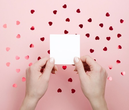 White sticker in female hands on pink background with hearts. Place to writeの写真素材
