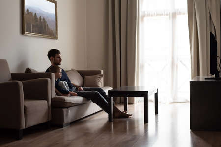 A young father and son are sitting on the sofa, watching TV. Joint leisure of father and son.の写真素材