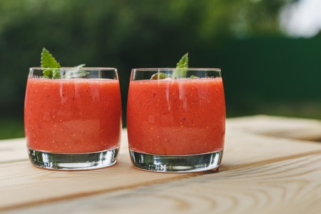 A glass of strawberry smoothie on a wooden table with mint in the garden