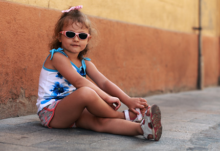 Fashion kid girl in sun glasses sitting on road near the wall and smilingの写真素材