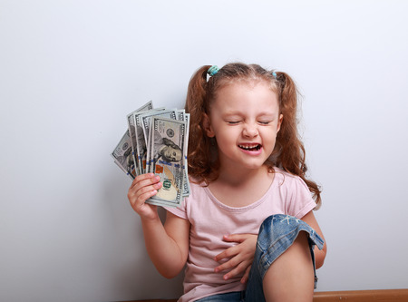 Laughing winner kid girl holding dollar with closed eye. Happy emotion child on blue backgroundの写真素材
