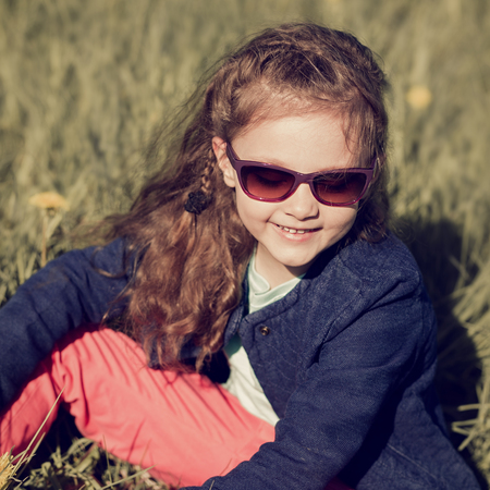 Fashion smiling kid girl sitting in green grass with flowers in fashion sunglasses and blue jacket. Toned closeup portraitの写真素材