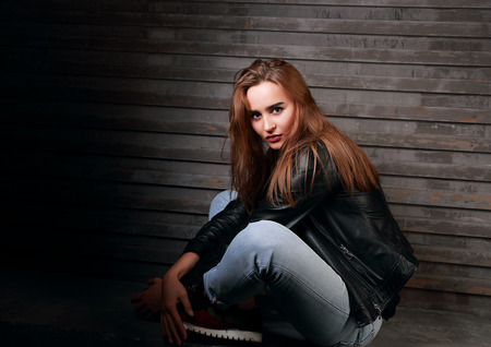 Beautiful red hair woman sitting on the stone in black jacket and looking on wall backgroundの写真素材