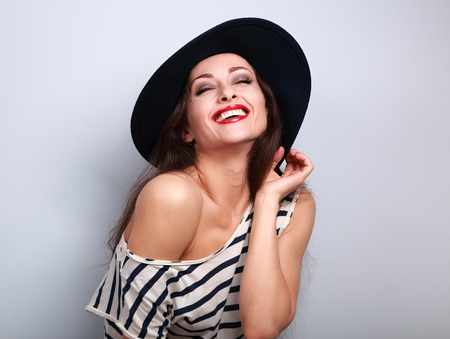 Happy toothy laughing brunette young woman in black hat with closed eyes on blue backgroundの写真素材